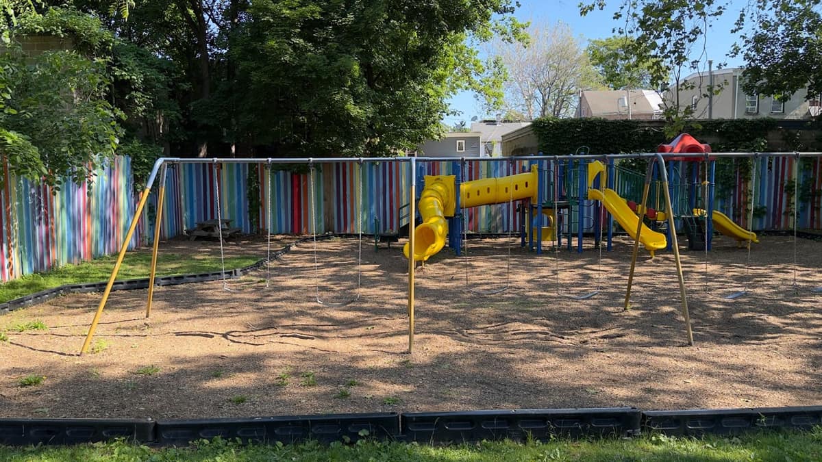 A yellow swing frame with empty chains hanging, in front of the play structure and rainbow-painted fence.