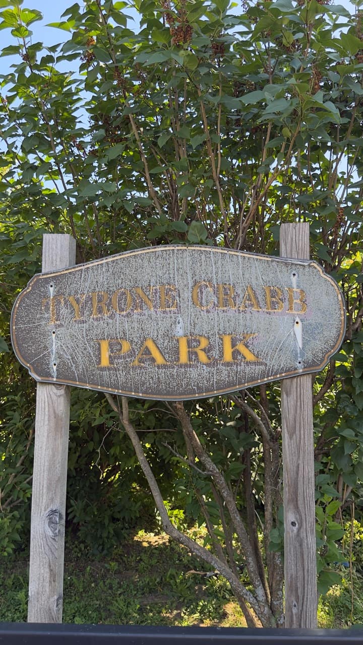 A faded silver and yellow Tyrone Crabb Park sign on two weathered wooden posts.