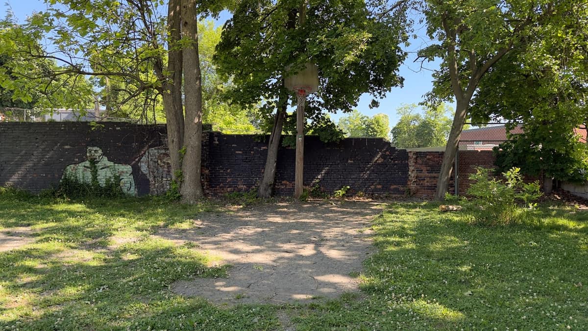 A dirt basketball area with a single hoop and a long brick wall behind, a faded figure painted on the wall.