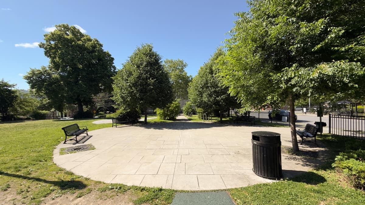 A wide concrete-paver plaza at Tyrone Crabb Park, ringed by benches and trees.