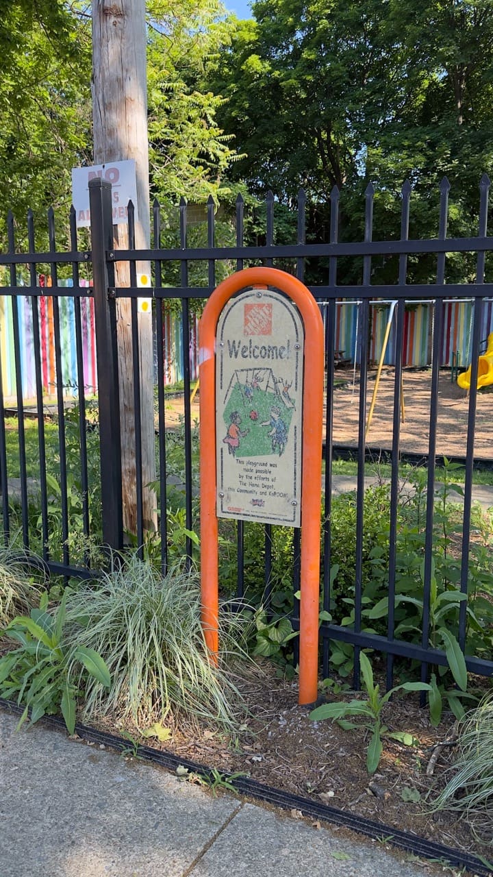 A weathered playground sign on a fence with sparse plantings in front, along a sidewalk.