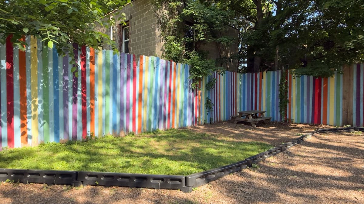 A brightly painted rainbow picket fence behind a wooden picnic table at Hasbrouck Street Park.