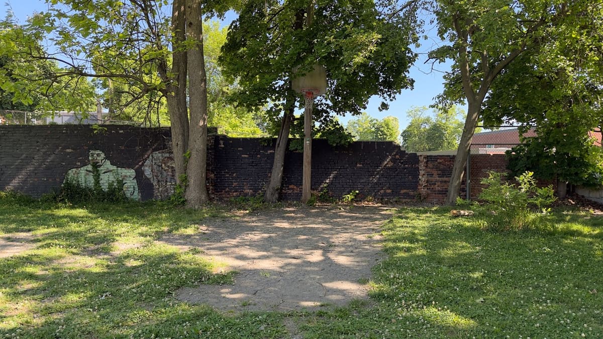 A dirt basketball area with a single hoop and a long brick wall behind, a faded figure painted on the wall.