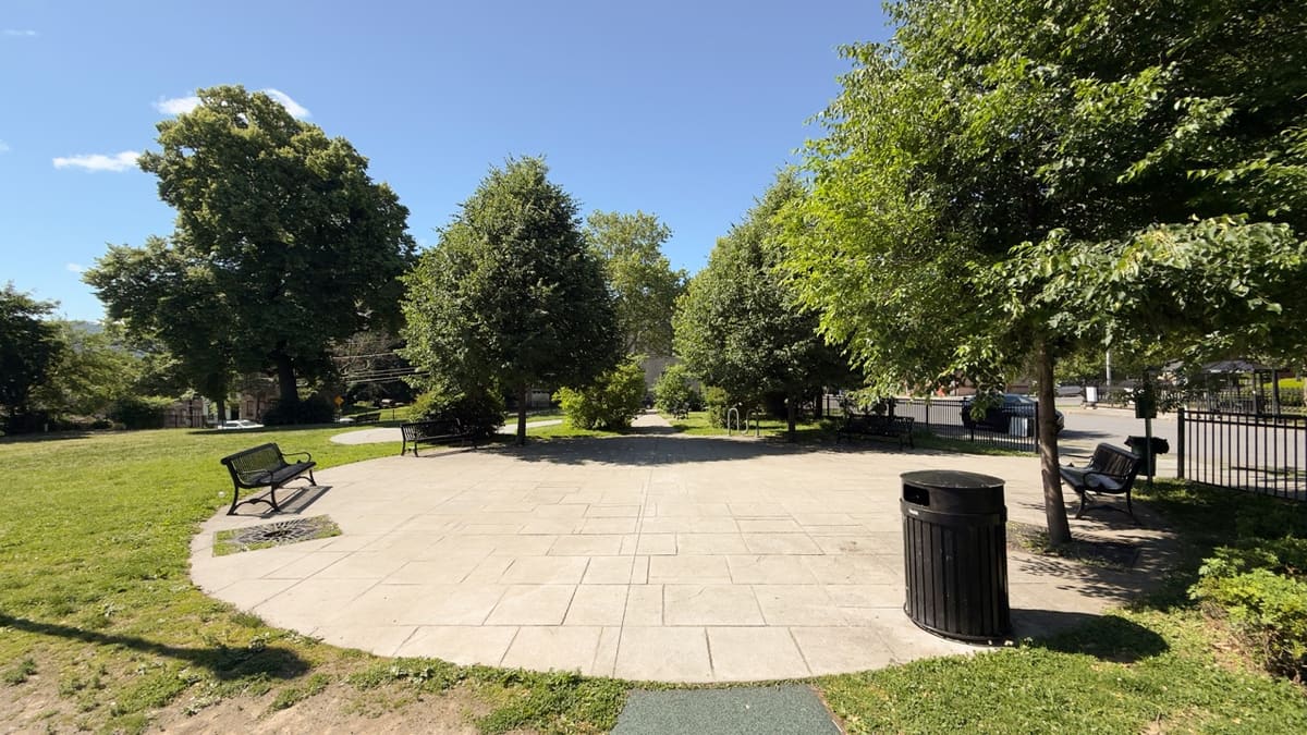 A wide concrete-paver plaza at Tyrone Crabb Park, ringed by benches and trees.