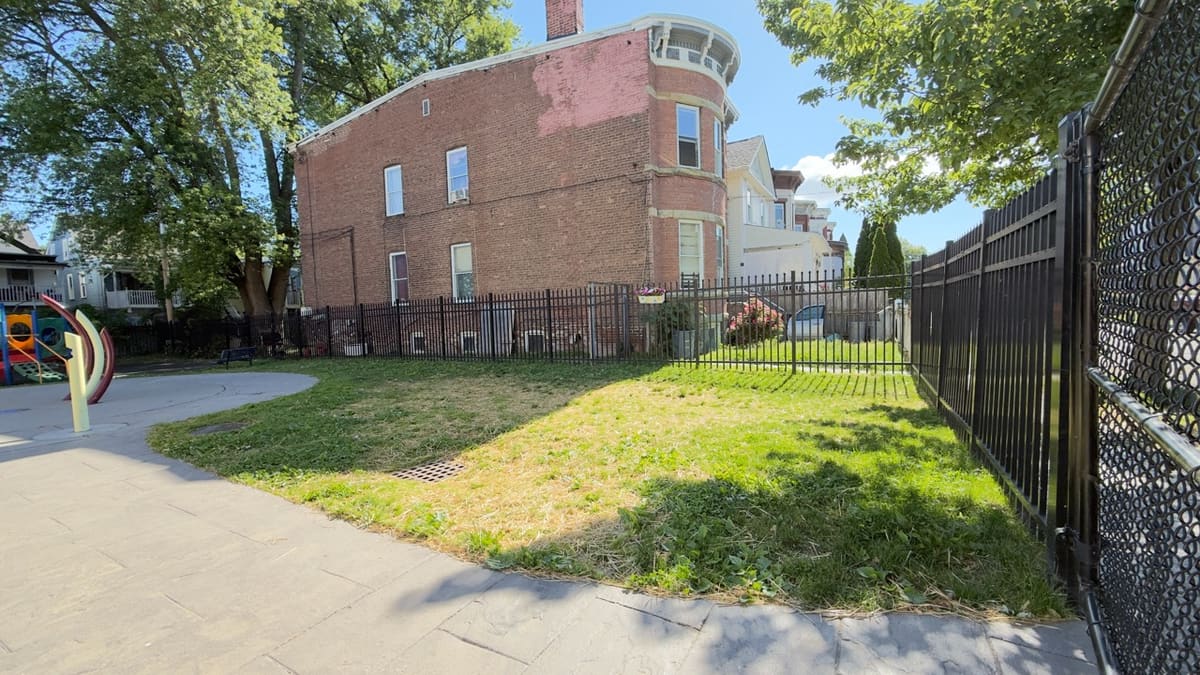 A grassy corner inside Xavier Lunan Park, with a brick building behind the fence and the play structure visible to the left.