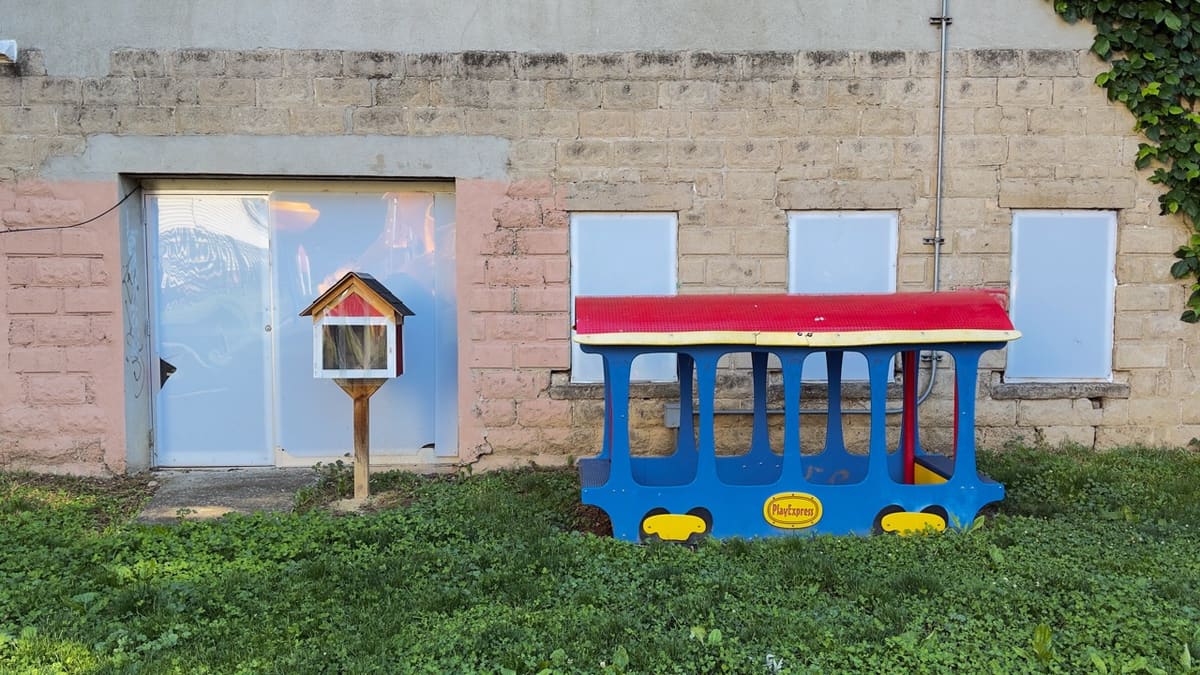 A small blue and red plastic play train beside a wooden Little Free Library against a cinder-block wall.